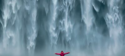 Iceland landscape photo of brave girl who proudly standing with his arms raised in front of water wall of mighty waterfall.